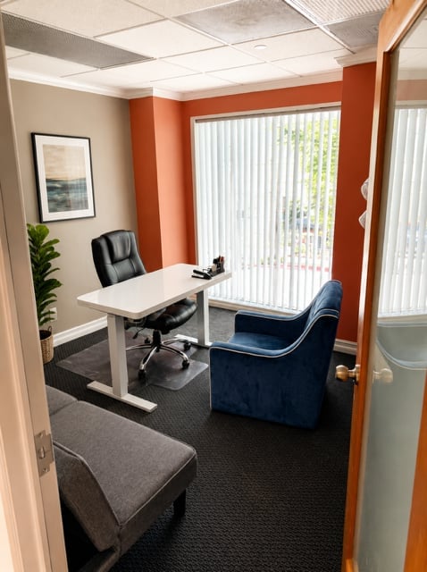 Modern office space with white desk, black chairs, orange accent walls, and natural light from vertical blinds